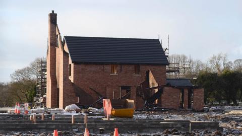 A red brick house with a black roof which is currently being built. There is the remains of a house in front of which appears to be burnt.