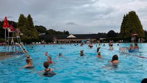 Swimmers in the outdoor pool watched over by a lifeguard sitting in a raised seat