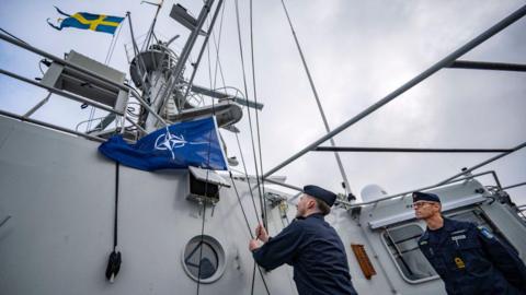 Two sailors on a military vessel. One of them is raising the NATO flag next to flag of Sweden while the other looks on. 