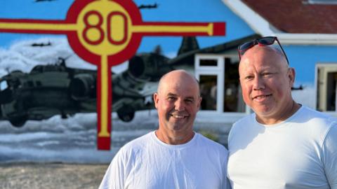 Two bald middle aged men smiling at the camera with a house in the background that has the red and yellow cross of the Guernsey flag with 80 in the middle and a large painting of an American B-17 bomber.