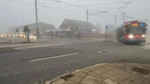 A red and blue supertram with Halfway as its destination is turning left across a junction. Cars are waiting across the road at traffic lights with their lights on due to the foggy conditions.