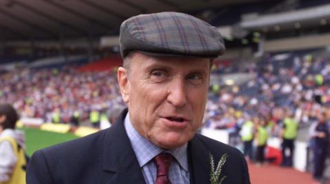 Hollywood actor Robert Duvall in a tartan bunnet in front of a big crowd. He is wearing a dark blue suit jacket, a red tie and shirt with some lucky heather in his buttonhole.