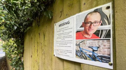 A missing poster for Martyn Richardson, which is pinned to a wooden fence. It has a picture of Mr Richardson, who is wearing black glasses and a red T-shirt. It also contains a picture of his bike, which is blue. 