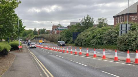 Riverside Road in Stockton is a four-lane road. There is a row of orange traffic cones in the middle, with a yellow diversion sign at the top. Cars are travelling in both directions.