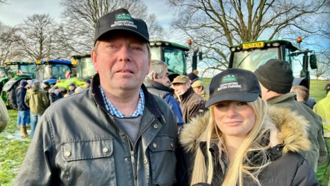 Dave Adamson with his daughter Sarah. Both are wearing baseball-style caps with the words: "Supporting British farming" on them. Sarah has long blonde hair and is wearing a winter coat. Dave is wearing a wax jacket. There are tractors in the background.