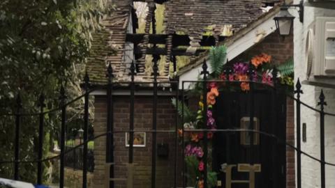 A restaurant with a timber roof and flowers on the front. The roof has been damaged by fire and is caged in, with trees visible through the gaps.