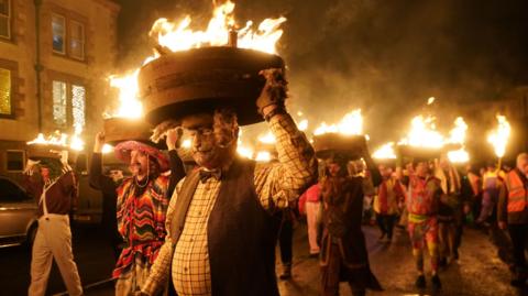 Men, known as guisers, carry burning whisky barrels on their heads through the streets during the Allendale Tar Barrels Parade. They are dressed in a variety of ways, some with colourful mexican ponchos and sombreros, others in white overalls and green wigs, or bow tie, gilet and animal mask.