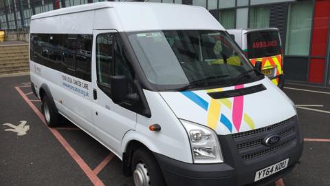 A white Ford Transit minibus with Wessex Cancer Support branding parked outside a hospital, with an ambulance beside it.