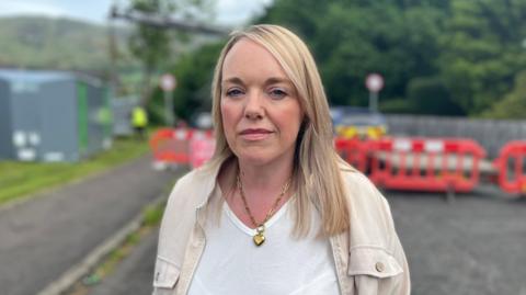 Ellen Forson, standing on a residential street in front of red safety barriers that block off the road, with trees and a hillside in the background. She is wearing a light-coloured jacket over a white top and a chain necklace, and is positioned in the foreground with the background softly out of focus.