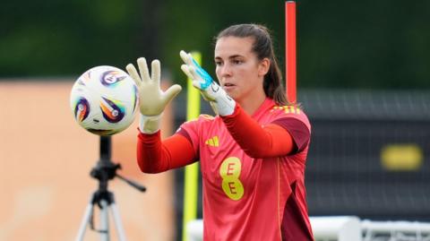 Olive Clark, with brown hair in a ponytail, has her arms out in front of her. She is about to catch a ball which is travelling towards her. She is wearing a red football shirt with EE in yellow letters. She is wearing white goalkeeper gloves. 
