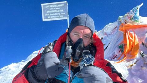 Man in breathing gear and very warm, padded clothing for mountain expedition gives the thumbs up- he is wearing very padded gloves, the sun is shining and the mountain and bits of material and a flag are seen behind him.