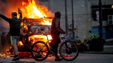 Two youths stand in front of a burning vehicle in Sunderland