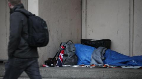 A homeless person sleeping rough in a blue sleeping bag a doorway. A man in a black coat, black backpack and dark blue jeans walks past.