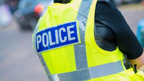 The back of a police officer, wearing a green high-visibility jacket with "police" written in blue and white and a black T-shirt inside.