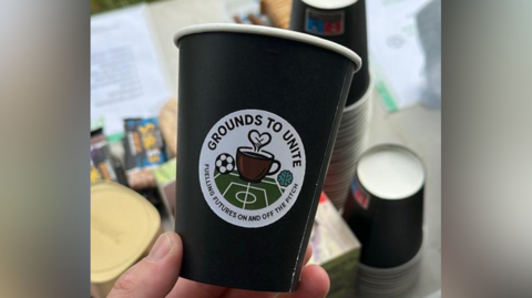 A black coffee cup with a white circular sticker on it that reads Grounds to Unite, fuelling futures on and off the pitch. A person is holding the club.