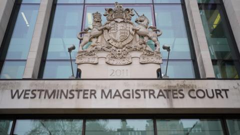 The entrance to Westminster Magistrates' Court. The name of the court is carved into grey stone and there is a crest for the court above the sign. Behind the sign are windows.