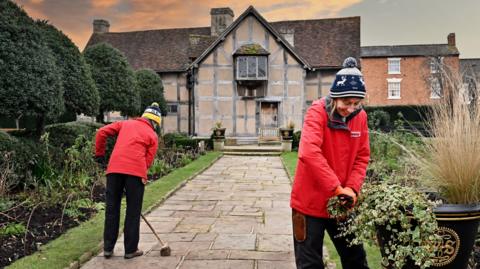 Two women wearing red jackets cleaning the front garden of a large half-timbered house.