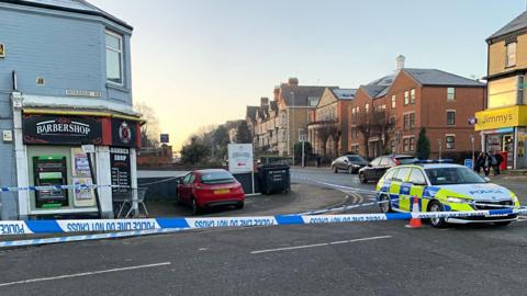 Police tape across a road, with a police car alongside. There is a barber's shop on a street corner to the left and a supermarket on the corner to the right. There are three-storey houses beyond the supermarket.