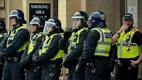 A line of six police officers in riot gear, including bright yellow vests and blue helmets with visors, look pensive as they stand shoulder-to-shoulder in front of a Victorian hotel building in Hull city centre