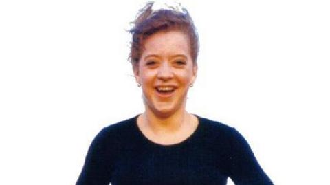 Vicky Glass smiling into the camera in a grainy photo. She has red hair tied up in a white scrunchie. She has small hooped earrings and is wearing a long sleeved black top.