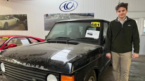 A man stands next to a black Range Rover. He is wearing tan trousers and a green half-zip fleece.