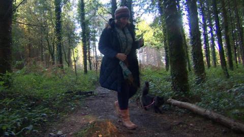 Helen Davies is walking Indie, a black cat, on a lead in woods. Ms Davies is wearing a woolly hat and scarf over a long coat.