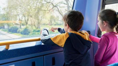 An image of a two children aged around 7 looking out of the front window on the upstairs of a bus. One has a brown ponytail and is wearing a pink long-sleeved top, the other has short brown hair and is wearing a navy jacket with yellow patches on the should and yellow netting in the hood.
The photo is taken from behind them