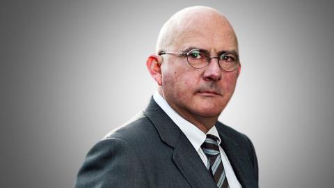A portrait of Andrew Norfolk wearing a suit and tie with glasses on looking directly at the camera against a grey background.