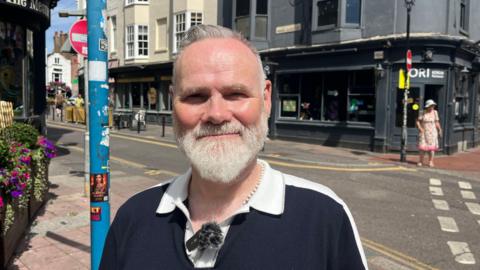 A man with white hair and beard is wearing a black shirt. He is standing on a busy street.