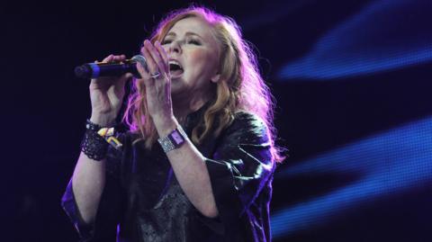 Carol Decker sings into a microphone at a concert, with purple light shining on her hair. She wars a black elbow-length gown and a diamante watch on one wrist.