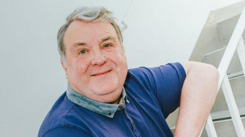 Russell Grant wearing a blue polo smiling at the camera and leaning on the handrail next to a set of stairs.