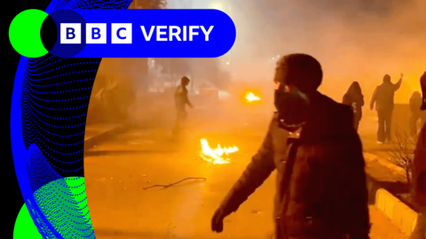 A protester wearing a mask walks on a highway in Iran. Fire, smoke and other protesters are visible in background.