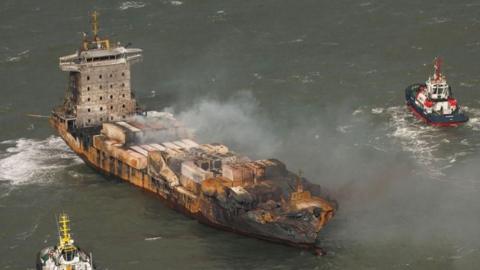 The Solong cargo ship with a fire-damaged hull and smoke rising from the deck, with two tug boats nearby,