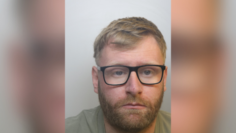 A custody image of Jack Wood, he has blonde hair, a blonde beard and black glasses. He is wearing a green tee-shirt.