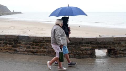 Two women walk along a beach promenade clutching an umbrella.
