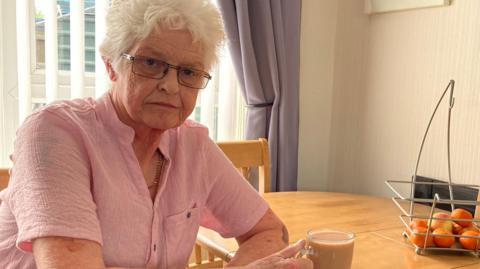 Sandra Robinson is wearing a pink shirt and is holding the handle of a mug which has a brown drink in it. On her dining room table is a fruit stand which has oranges inside.