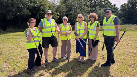 A group of volunteers stood in the park holding their garden tools and wearing high vis jackets.