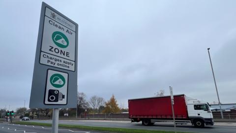 A road sign indicates Bradford's Clean Air Zone. It is captioned "Charges apply, pay online". A red lorry drives along the dual carriageway in the background.