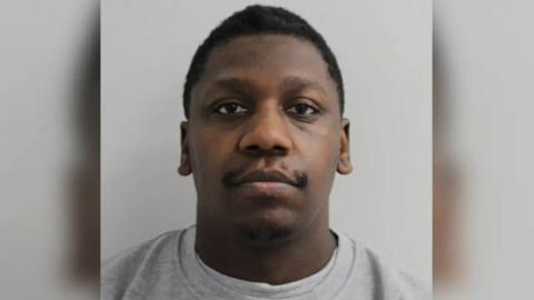 A mugshot style image of a man. He has a blank expression and is staring down the camera. He is wearing a plain grey jumper and has a moustache and short hair