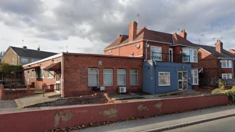 A street view image of the red brick building. The site looks empty and unused, paint is peeling off the wall in front of it. 