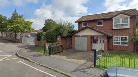 A brick house estimated to date to the 1980s, with white PVC doors and windows and a grey garage door. There are metal railings around the property. To the left, the entrance gates to a school car park are visible.