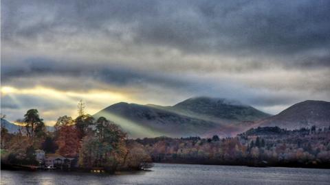 The sun tries to squeeze through a break in the cloudy sky sending rays over mountains and trees 