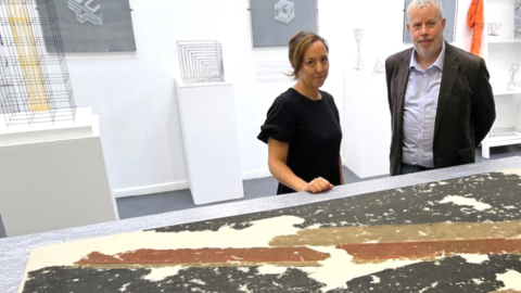 Dr Howard Fuller is wearing a suit with a shirt and is standing to the right, as you look at the image ,of Sarah Morris. She is wearing a black top and has brown hair, tied back. The flag is partially seen on a table in front of them and slightly resembles a simpler Union Jack design.