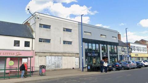 A weathered white building with several small windows and a boarded-up ground floor is joined to a modern-looking grey shopfront with large glass windows and signage, followed by additional businesses. Several cars are parked along the street, while a few pedestrians walk along the pavement. The road has marked lanes and parking areas, and tall blue streetlights line the pavement. Above, the scene is set against a bright blue sky with scattered white clouds.