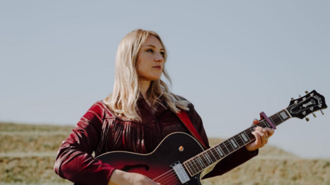 Dolly Mavies in a dark red jacket standing in a field holding a guitar