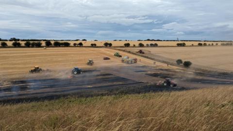 A partially charred field can be seen down a gentle slope, with long grass in the foreground of the photo. Smoke is rising from the blackened ground, and there are six tractors around or on it. There are also two fire engines and a car parked on the field. There is a hedge and another untouched field to the right of the image.