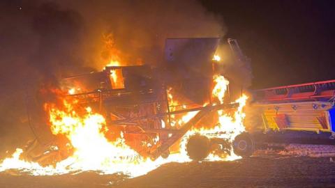 A combine harvester is on fire, it is night time, flames engulf the machine. It is in a field. 
