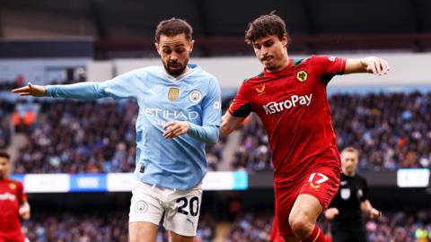 Hugo Bueno, wearing Wolves' red away kit, challenges Manchester City's Bernardo Silva who is on his right. Bueno has one arm behind Silva's back and they are both looking down at the ball, which is not in shot. 