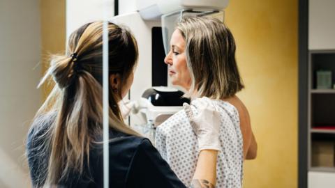 A woman stands in a hospital gown undergoing a mammogram. A medical practitioner stands beside her in a black top.