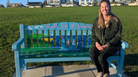 Helena Chessher sitting on the right end of a colourful bench painted with floral and coastal designs